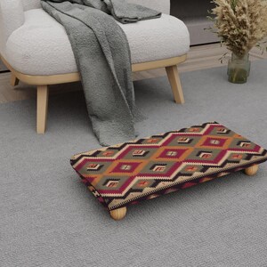 Peut inclure: Un repose-pieds rectangulaire avec un motif géométrique aux tons de rouge, orange, vert et beige. Le repose-pieds a quatre pieds courts en bois arrondis. Il est posé sur un tapis gris près d'un fauteuil blanc et d'une couverture grise.