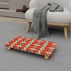 Puede incluir: Un reposapiés rectangular con un patrón ovalado rojo y blanco y detalles de diamantes azules. El reposapiés tiene cuatro patas de madera y está colocado sobre una alfombra gris. El diseño es de estilo retro.