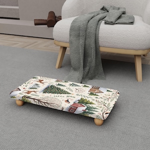 May include: A rectangular footstool with a winter scene pattern featuring deer, trees, and houses. The stool has a cream-colored background and is supported by four small, rounded wooden legs. The design includes shades of green, brown, and red.