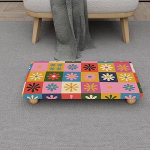 May include: A rectangular footstool with a colorful floral pattern in a grid design. The flowers are in various colors, including pink, yellow, blue, and black, set against a background of different colors. The footstool has four wooden legs.