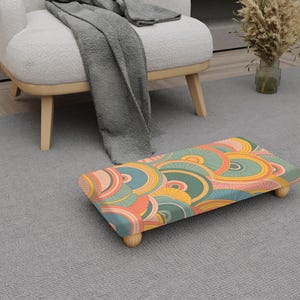 May include: A rectangular footstool with a colourful retro design featuring orange, yellow, teal, and green concentric circles. The footstool has four wooden legs and is placed on a grey carpet.