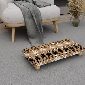May include: A rectangular footstool featuring a repeating pattern of black, white, and tan leaf and geometric designs on a tan background. The footstool has four wooden legs and is on a grey rug.