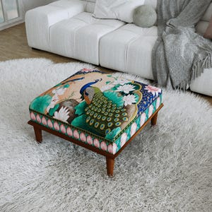 May include: A square ottoman featuring a vibrant peacock and lotus flower print on the top surface. The sides are decorated with a repeating pink lotus petal pattern. The piece is supported by four short, dark brown wooden legs.