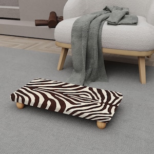 Op de afbeelding: Een rechthoekig dierenbed met een zebraprint in bruin en wit. Het bed staat op vier kleine, afgeronde houten poten. Het ligt op een grijs tapijt, dichtbij een witte fauteuil met een grijze deken.