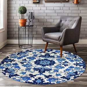 Op de afbeelding: Een rond vloerkleed met een witte achtergrond en een herhalend blauw bloemen- en plantenpatroon. Het kleed ligt op een donkere houten vloer voor een grijze gestoffeerde fauteuil met knoopdetails en lichtbruine houten poten.