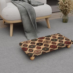 Peut inclure: Un repose-pieds rectangulaire avec un fond beige et un motif répétitif de formes géométriques rouges, vertes et marron. Le repose-pieds a quatre pieds en bois et est placé sur un tapis gris près d'un fauteuil blanc.