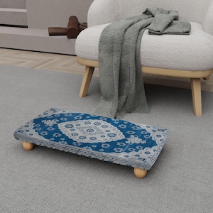 May include: A rectangular footstool with a blue and white patterned top, supported by four small, rounded wooden legs. The footstool is placed on a gray carpet, with a white armchair and a gray blanket in the background.