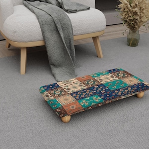 May include: A rectangular footstool with a patchwork design in shades of blue, teal, orange, and beige. The footstool has four short, rounded wooden legs. The design features floral and geometric patterns.