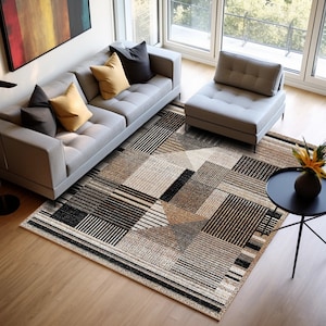 Op de afbeelding: Een grijze bank met gele en zwarte kussens staat op een beige en zwart geometrisch tapijt. Een grijze poef en een kleine ronde tafel met een vaas met bloemen staan ook op het tapijt.