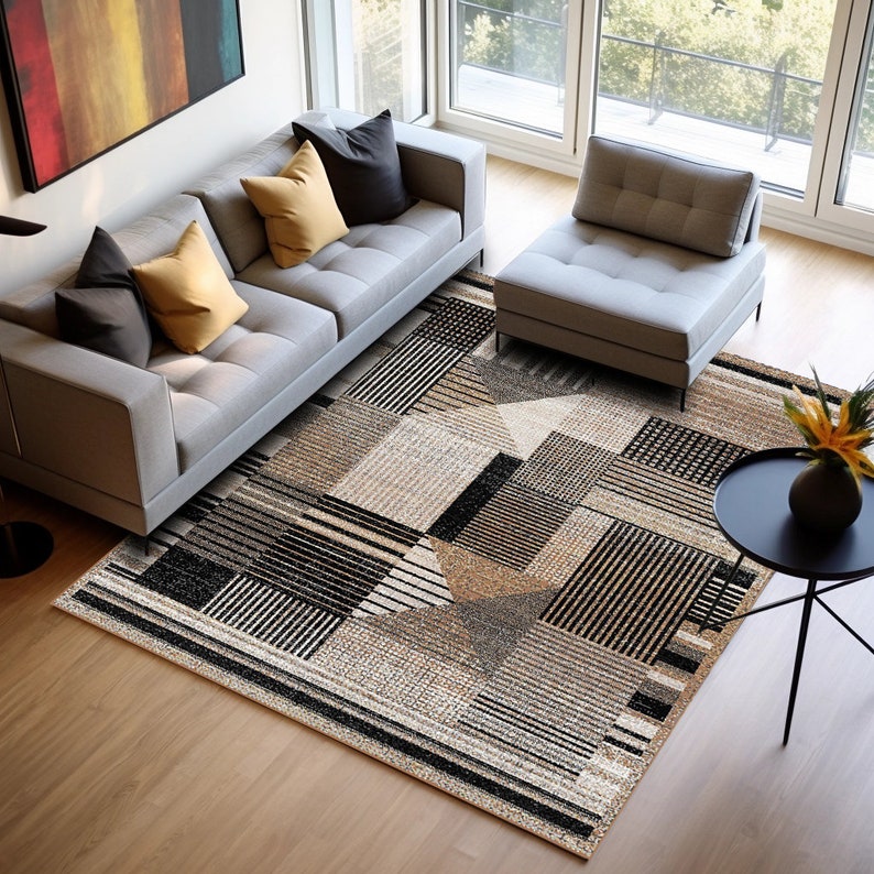 Op de afbeelding: Een grijze bank met gele en zwarte kussens staat op een beige en zwart geometrisch tapijt. Een grijze poef en een kleine ronde tafel met een vaas met bloemen staan ook op het tapijt.