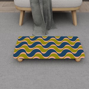 Puede incluir: Un reposapiés decorativo con un patrón ondulado en verde oliva, azul marino y rosa coral. El taburete rectangular está sostenido por cuatro pequeñas patas de madera. El diseño es de estilo retro, de mediados de siglo.