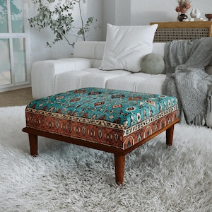 a cozy living room with a plush white sofa, a colorful patterned ottoman, and a large window with white curtains. The room has a warm and inviting atmosphere, with a rug and a plant adding to the homely ambiance.