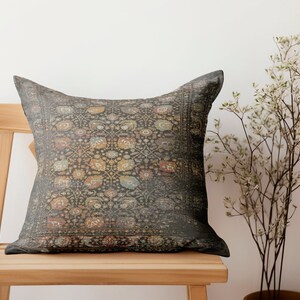 K&ouml;nnte beinhalten: Ein quadratisches Zierkissen mit einem dunkelgrauen, abgenutzten Muster im Vintage-Stil. Das Kissen liegt auf einem hellen Holzstuhl. Das Muster umfasst florale und geometrische Elemente in Orange-, Blau- und Gelbt&ouml;nen.