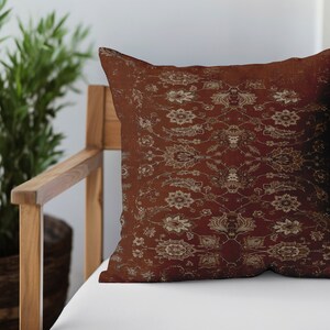 May include: A decorative throw pillow with a red and gold floral pattern. The pillow is sitting on a wooden chair with a white cushion.
