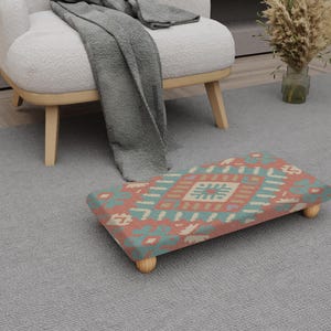 May include: A rectangular footstool with a colourful geometric pattern in shades of coral, teal, and cream. The footstool has four short, rounded wooden legs. The design is reminiscent of a traditional rug.