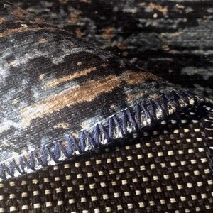 May include: Close-up of a dark blue rug with a subtle pattern of brown and white. The rug has a dark blue stitched edge and a black and white checkered backing.