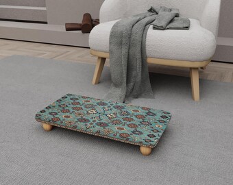Op de afbeelding: Een rechthoekige voetenbank met een turquoise stoffen bovenkant met bloemen- en geometrische patronen. De voetenbank heeft een houten frame en vier afgeronde houten poten. De voetenbank staat op een grijs tapijt.
