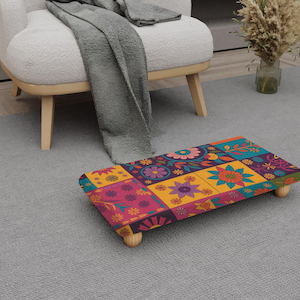 May include: A rectangular footstool with a colorful floral patchwork design. The footstool is supported by four small, rounded wooden legs. The color palette includes pink, yellow, blue, and purple hues, creating a vibrant and decorative piece.