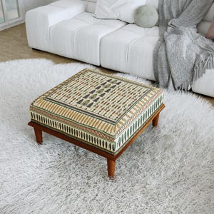 Op de afbeelding: Een vierkante poef met een crèmekleurige stoffen bekleding voorzien van een herhalend patroon van groene en bruine botanische motieven. De poef rust op vier korte, donkerbruine houten poten en heeft een gestructureerd ontwerp.