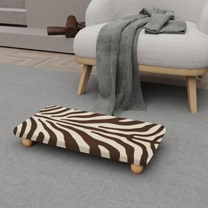 May include: A rectangular footstool featuring a zebra print in brown and cream. It has rounded wooden legs and sits on a grey carpet. The background includes a white armchair and a grey blanket.