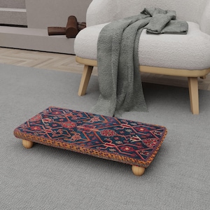 May include: A rectangular footstool with a patterned design in shades of blue, red, and gold. The footstool has rounded wooden legs and a decorative border. The footstool is placed on a gray carpet, near a white armchair.