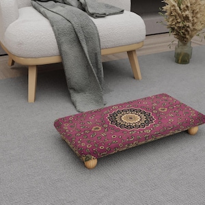 May include: A rectangular footstool with a magenta and gold patterned top, featuring a central medallion design. The footstool rests on four short, light-coloured wooden legs. The footstool is placed on a grey carpet.