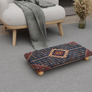 May include: A rectangular footstool with a patterned design in shades of blue, red, and beige. The footstool has a wooden frame and four short, rounded legs. It is placed on a gray carpet near a white armchair.
