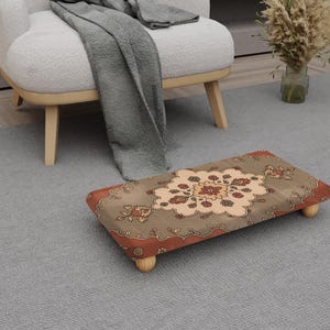May include: A rectangular footstool with a patterned top featuring a central floral design in beige, red, and brown. The footstool has a wooden frame and short, rounded legs. The footstool is placed on a gray rug.