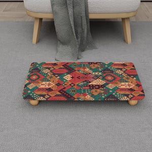 May include: A rectangular footstool with a colorful geometric pattern in shades of red, green, and orange. The footstool is supported by four small, round wooden legs. The design features diamond and floral shapes.