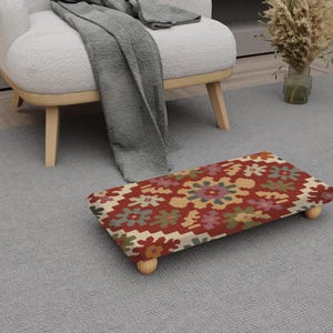 May include: A rectangular footstool with a patterned fabric top in shades of red, beige, green, and purple. The footstool has four short, rounded wooden legs. The footstool is placed on a grey carpet near a white armchair.