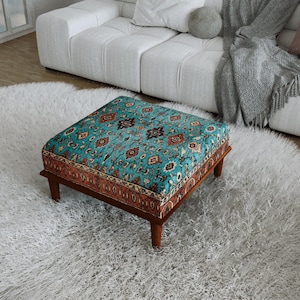 May include: A square ottoman featuring a vibrant turquoise fabric top with a detailed multi-colored floral and geometric pattern. The sides are accented with a rust-colored border featuring intricate designs, supported by four dark wooden legs.
