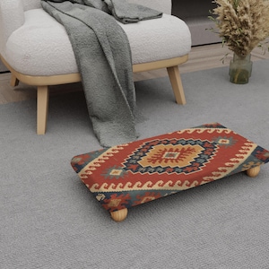 May include: A rectangular footstool with a patterned rug design in red, blue, and yellow. The footstool has four wooden legs and is placed on a grey carpet. A white armchair and a grey blanket are in the background.