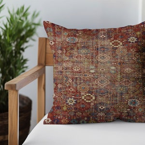 以下が含まれることがあります： ヴィンテージラグのデザインの装飾用スロー枕。枕は赤、青、ベージュの色合いで複雑なパターンが特徴です。木製の椅子に白いクッションの上に置かれ、背景には緑の植物があります。