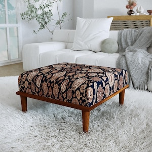 May include: A square ottoman featuring a dark navy blue fabric upholstery with a dense, intricate floral and paisley pattern in shades of tan and cream. The piece is supported by four tapered wooden legs with a warm, medium-brown finish.