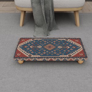 May include: A rectangular footstool with a patterned rug design. The rug features a central diamond pattern in shades of blue, red, and beige. The footstool rests on four small, rounded wooden legs, and is placed on a gray carpet.