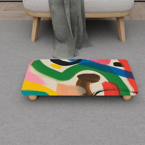 Peut inclure: Un repose-pieds rectangulaire avec un motif abstrait coloré. Le dessus présente des coups de pinceau verts, bleus, roses, jaunes et bruns sur un fond crème. Il est soutenu par quatre petits pieds ronds en bois.