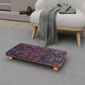May include: A rectangular footstool with a patterned rug top in shades of blue, red, and orange. The footstool has four rounded wooden legs. The rug design features floral and geometric motifs.