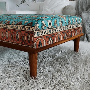 a modern, wooden ottoman with a vibrant, patterned cushion on top, placed on a fluffy white rug.