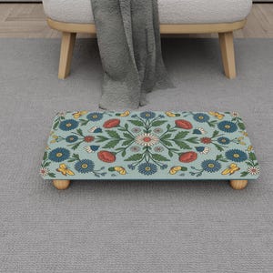Puede incluir: Un reposapiés rectangular con un estampado floral en tonos azules, rojos, amarillos y blancos. El reposapiés tiene un fondo azul claro y está sostenido por cuatro patas de madera pequeñas y redondeadas. El diseño presenta motivos florales y de hojas simétricos.