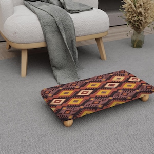 Puede incluir: Un reposapiés rectangular con un colorido patrón geométrico en tonos rojos, amarillos y marrones. El reposapiés tiene cuatro patas de madera y está colocado sobre una alfombra gris. El diseño recuerda al arte del suroeste o tribal.