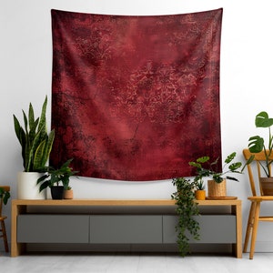 May include: A red tapestry with a distressed, textured look hangs on a white wall above a gray entertainment center. The tapestry has a subtle pattern of gold swirls and lines. The entertainment center is flanked by two wooden chairs and several potted plants.