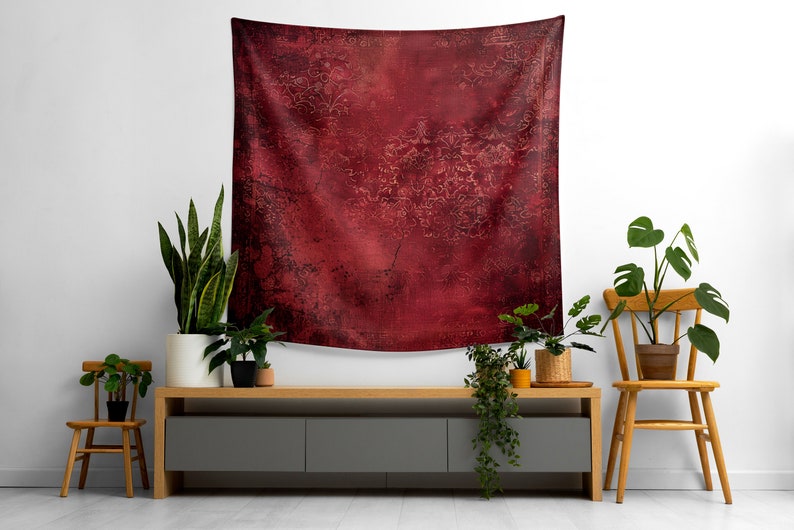 May include: A red tapestry with a distressed, textured look hangs on a white wall above a gray entertainment center. The tapestry has a subtle pattern of gold swirls and lines. The entertainment center is flanked by two wooden chairs and several potted plants.