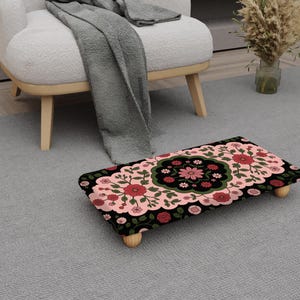 May include: A rectangular footstool with a floral pattern in red, pink, and green on a black background. The footstool has four wooden legs and is placed on a gray rug. The design features a central floral medallion.
