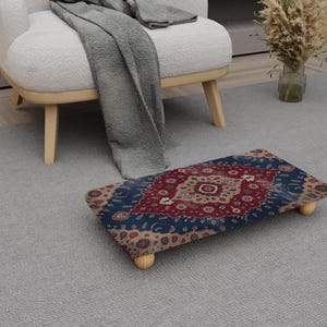 May include: A rectangular footstool with a patterned design in shades of red, blue, and beige. The footstool has four short, rounded wooden legs. The design features a central medallion and floral motifs.