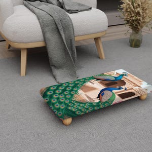 May include: A rectangular footstool featuring a peacock feather design and two peacock illustrations. The footstool has wooden legs and is placed on a grey carpet. The peacock feathers are green, blue, and gold.