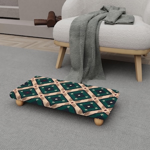 Op de afbeelding: Een rechthoekige voetenbank met een groen, beige en rood ruitpatroon. De voetenbank heeft vier houten poten. De voetenbank staat op een grijs tapijt, in de buurt van een witte fauteuil met een grijze deken.