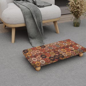 May include: A patterned footstool with a geometric design in red, orange, and brown. It has four small wooden legs. The footstool is on a grey carpet, near a white armchair. The footstool measures approximately 40 cm in length.