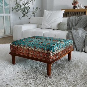 a cozy living room with a plush white sofa, a colorful patterned ottoman, and a large window with white curtains. The room has a warm and inviting atmosphere, with a rug and a plant adding to the homely ambiance.