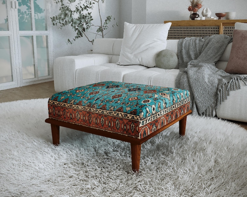 a cozy living room with a plush white sofa, a colorful patterned ottoman, and a large window with white curtains. The room has a warm and inviting atmosphere, with a rug and a plant adding to the homely ambiance.