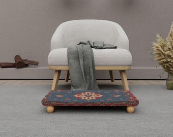 Op de afbeelding: Een kleine, rechthoekige voetenbank met een patroon van een tapijt in tinten blauw, rood en crème. De voetenbank rust op vier afgeronde houten poten. Een witte gestoffeerde stoel en een grijze deken staan op de achtergrond.